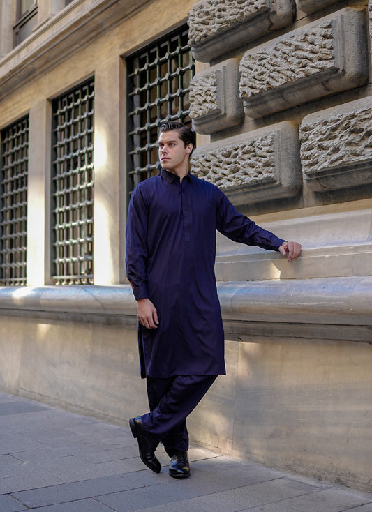 A man standing on the sidewalk wearing a navy blue, unstitched, solid-patterned fabric. Sheikh Gulzar Fabrics Bosphorus Blue — premium unstitched navy-blue fabric styled on model, luxury men’s shalwar kameez fabric Pakistan.