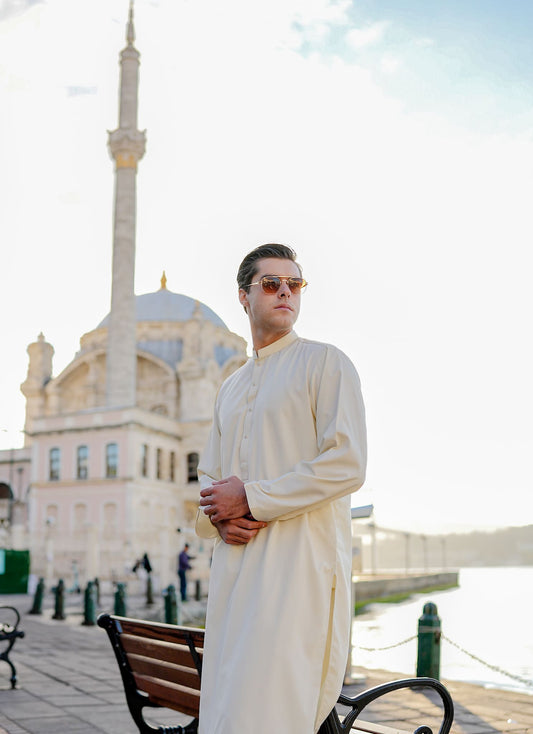 Person in a white outfit standing in front of a mosque with a minaret. Sheikh Gulzar Fabrics Ephesus Cream — premium unstitched creamy-white fabric styled on model in mosque courtyard, luxury men’s shalwar kameez fabric Pakistan.