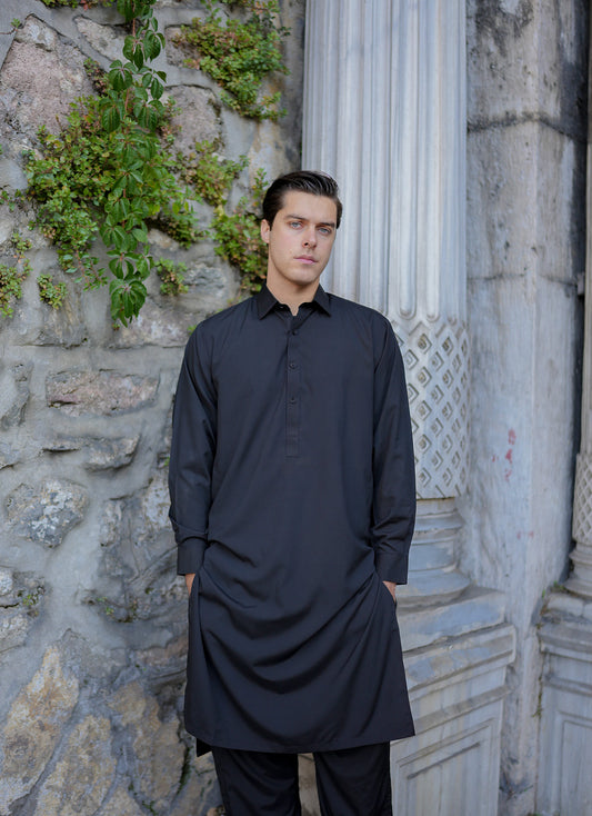 A man standing in front of a columned building, wearing a black traditional outfit with a slight shine, looking off to the side. Sheikh Gulzar Fabrics Istanbul Noir — elegant black fabric styled on model outdoors, premium unstitched shalwar kameez fabric Pakistan.