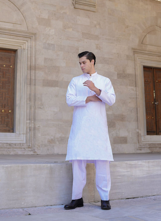 A man standing with one hand against a wall, wearing a white sherwani suit with a dupatta, in a columned hallway with intricate ceiling details. Sheikh Gulzar Fabrics White Sultan fabric — premium white unstitched shalwar kameez fabric styled in luxury sherwani, breathable and elegant men’s fabric Pakistan.